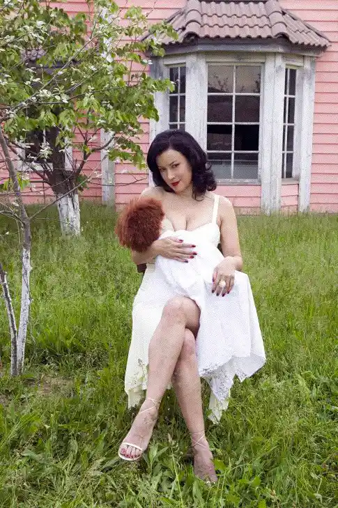 A woman in a white dress sits on grass in front of a pink house, holding a small reddish-brown dog. She looks directly at…