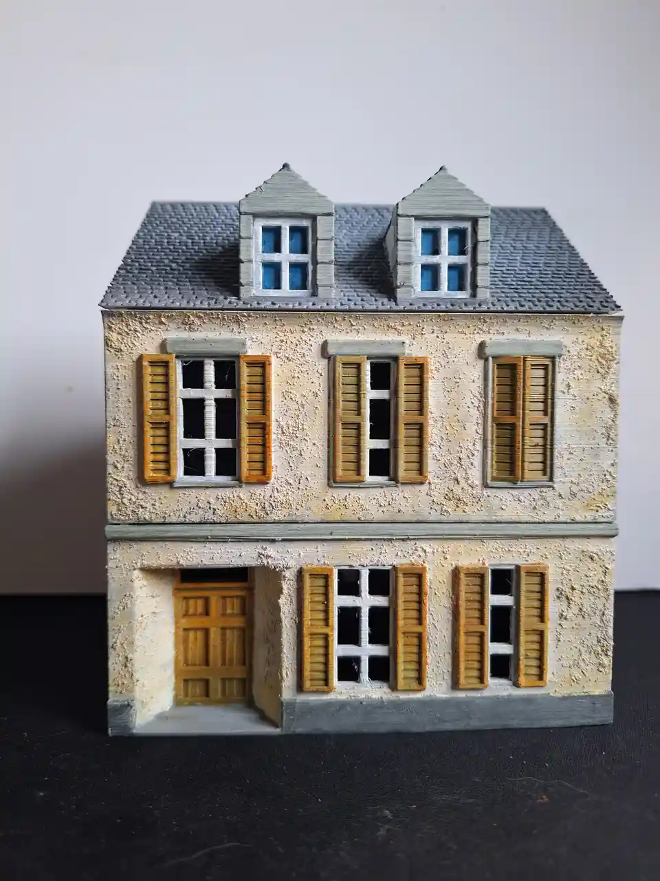 A detailed miniature model of a two-story house with a gray roof, dormer windows, and yellow shutters, placed on a black…