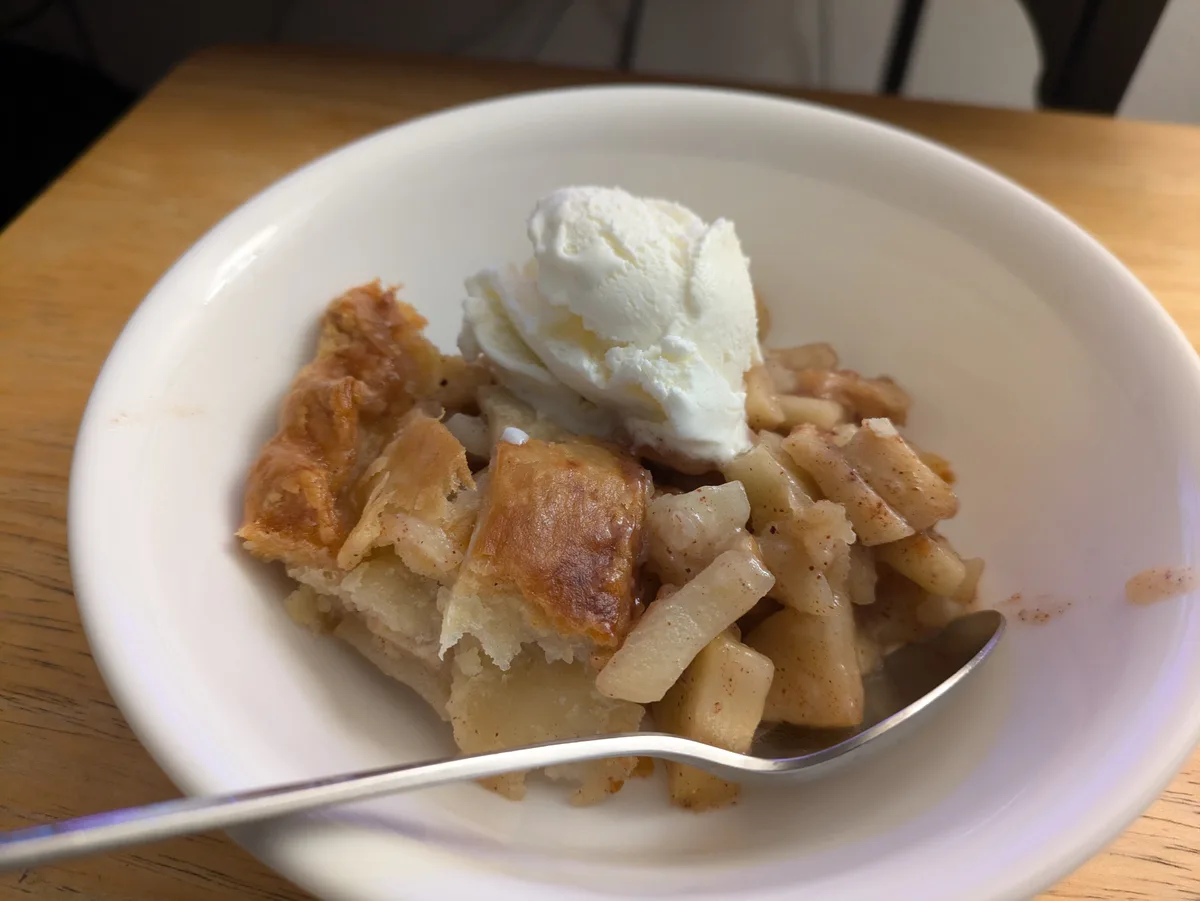 A close-up photo shows a white bowl filled with a serving of apple pie, featuring golden-brown pastry, cooked apple chunks…