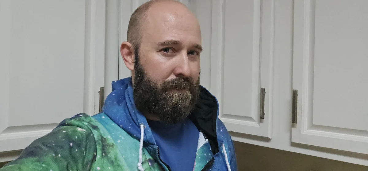 A bald man with a full beard wearing a blue and green galaxy-print hoodie stands in front of white kitchen cabinets. He…