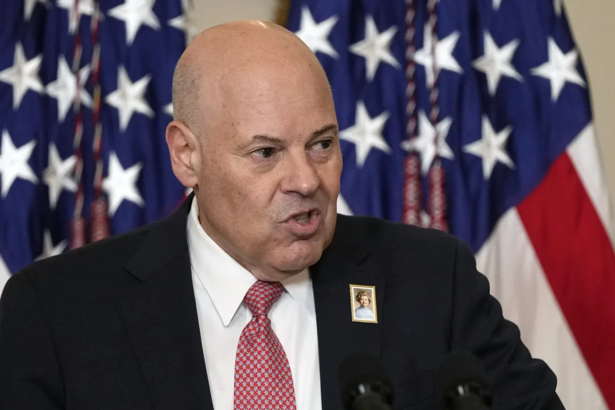 a bald man in a dark suit and red tie speaks at a podium with american flags behind him, wearing a small pin of a woman's…