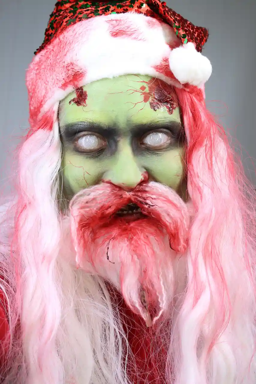 A person dressed as a zombie Santa Claus with green face paint, bloody wounds, and a pink and white wig, wearing a red…