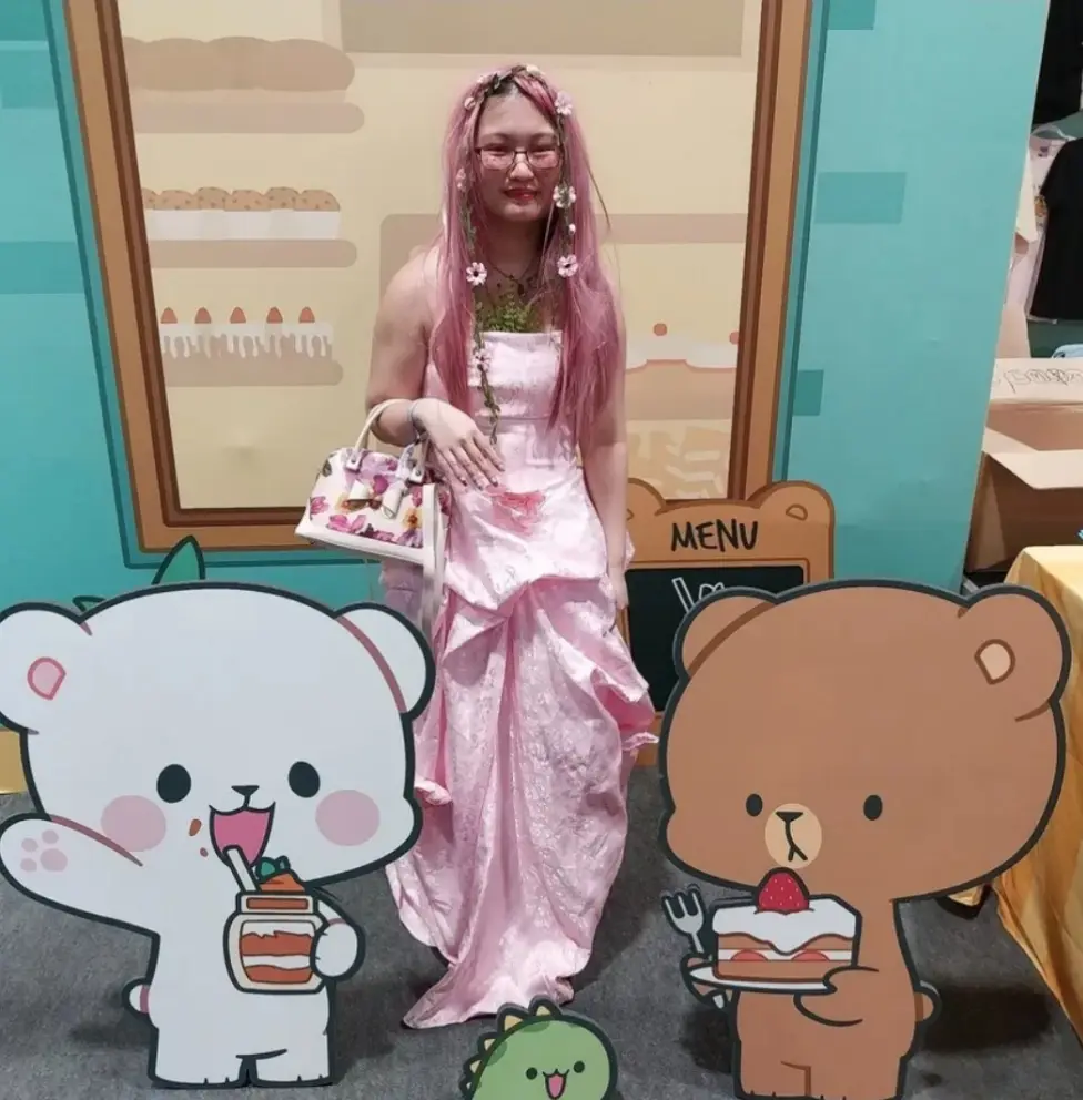 A person with pink hair and glasses, wearing a pink dress and holding a floral handbag, stands between two large cartoon…