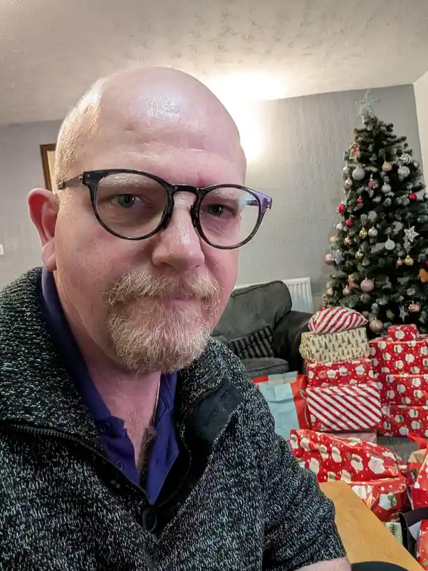 A bald man with glasses and a goatee stares directly at the camera with a weary expression. Behind him, a decorated…