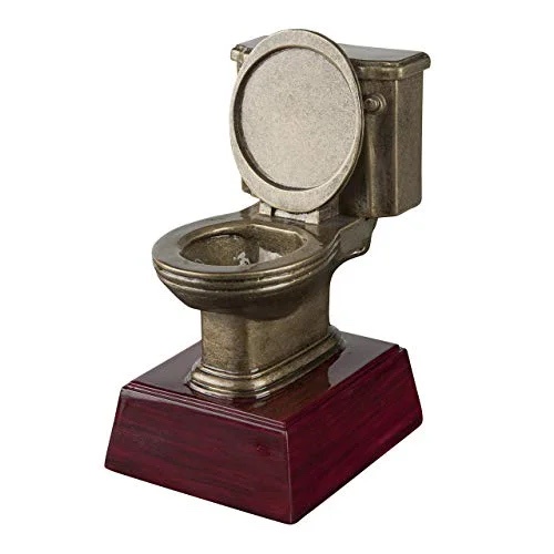 A small, ornate, bronze-colored toilet figurine sits on a polished dark red wooden base, with the toilet lid open. The…