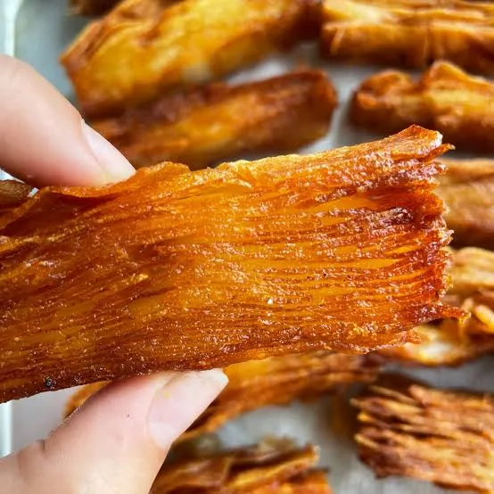 A close-up photo shows a hand holding a thick, layered, golden-brown snack that looks like crispy fried plantain or a…