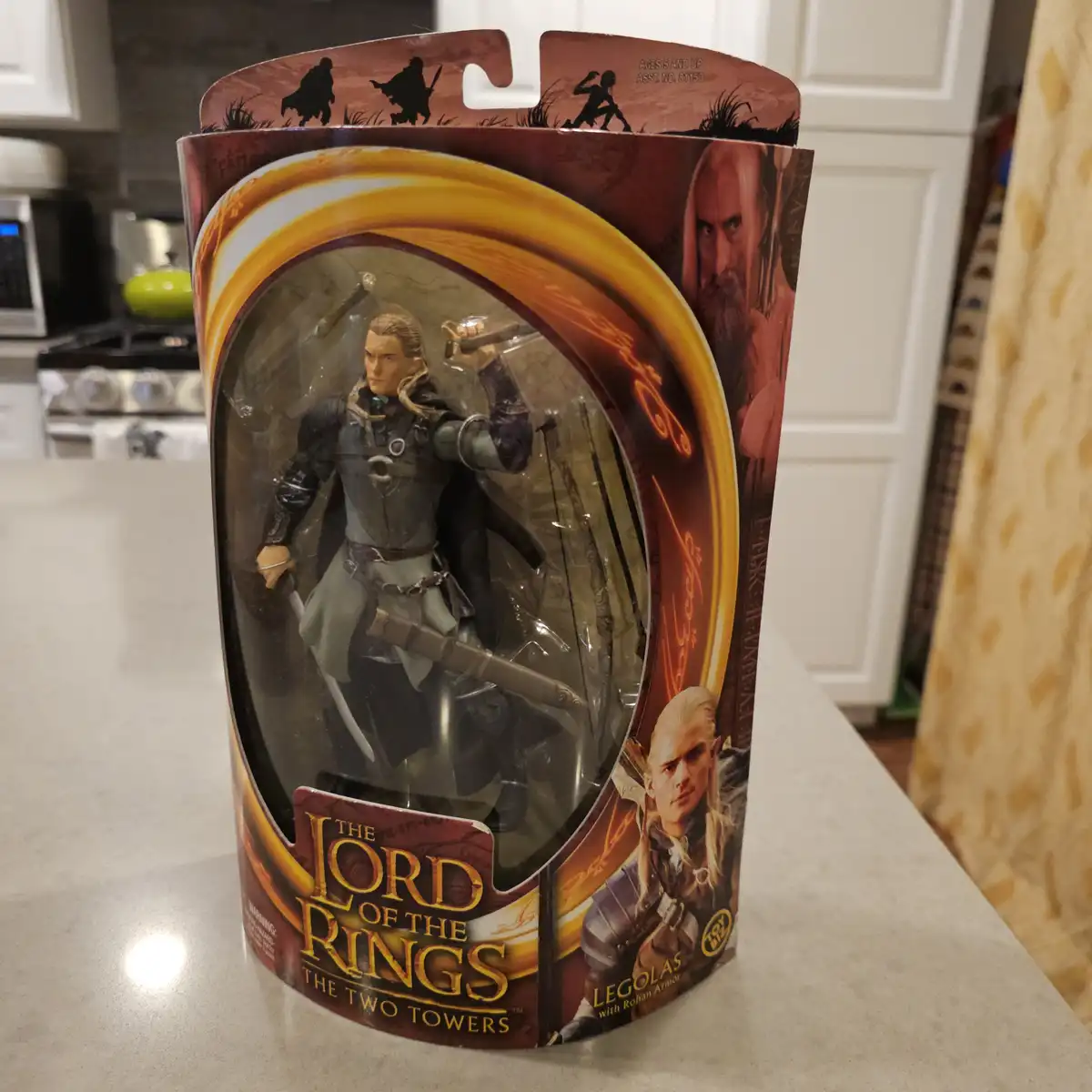 A Lord of the Rings action figure of Legolas, packaged in its original Toy Biz box, is shown standing on a kitchen counter…