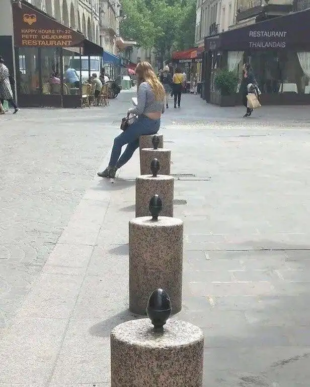 A woman with long blonde hair sits casually on a row of stone bollards in a pedestrian street, wearing a grey crop top and…