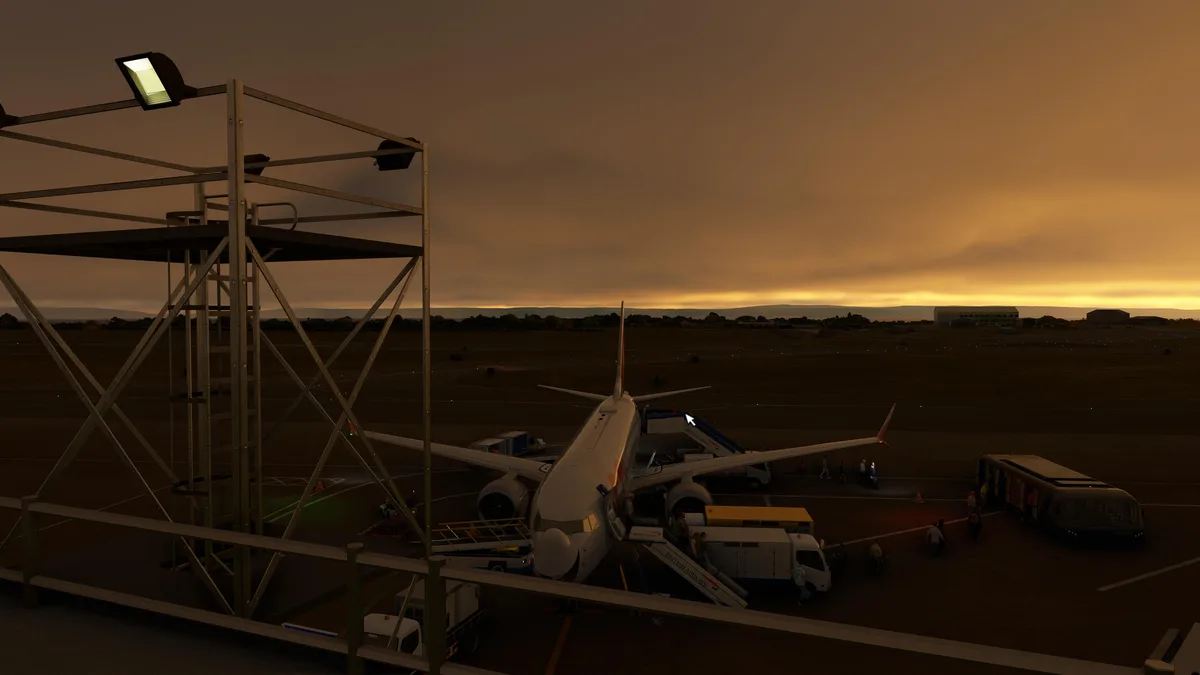 An airport tarmac scene at dusk with a British Airways plane parked at a gate, surrounded by ground crew, baggage carts, and…