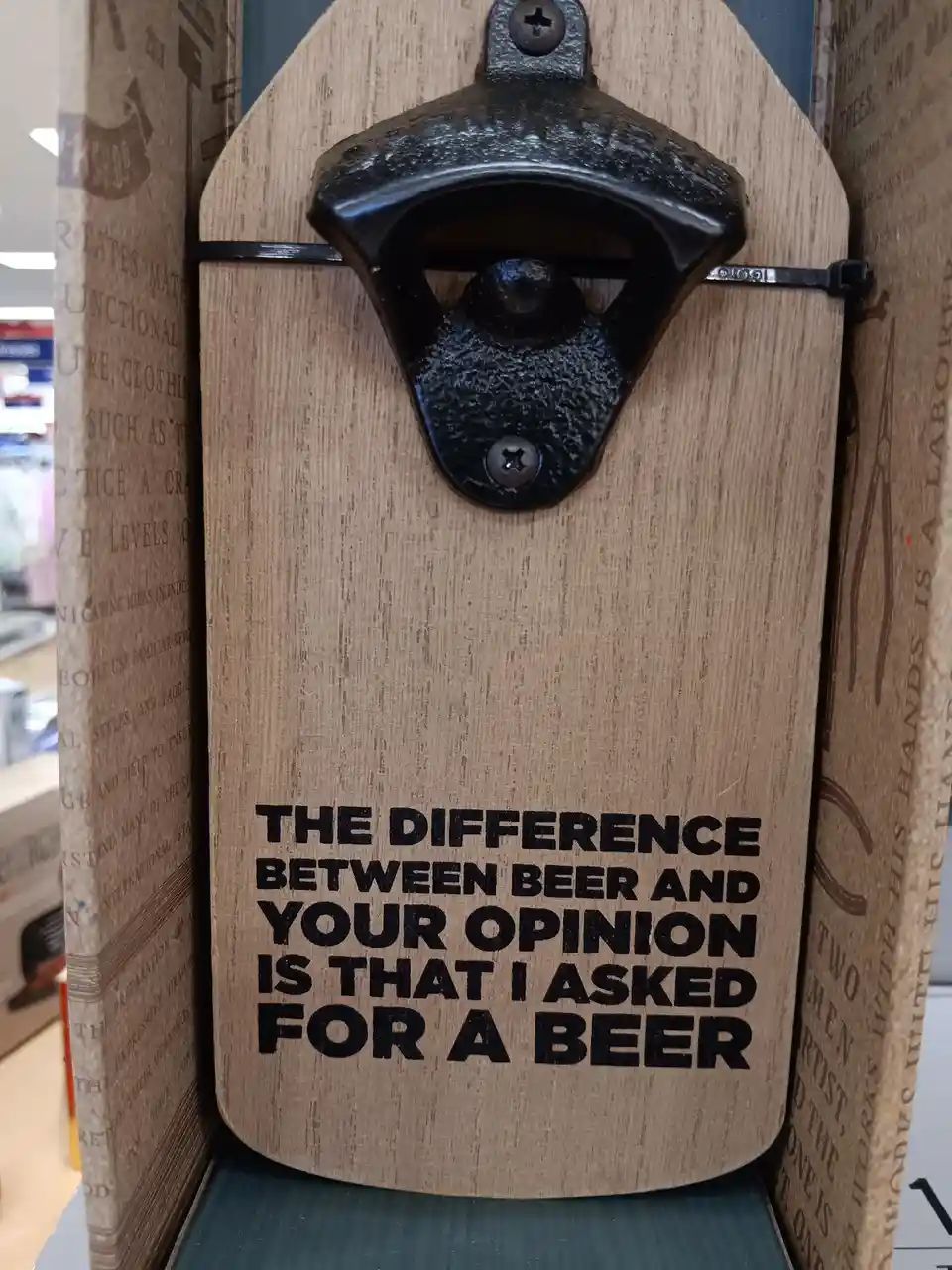 A wooden wall-mounted bottle opener with black metal hardware is displayed in a store box. The wood has bold black text that…