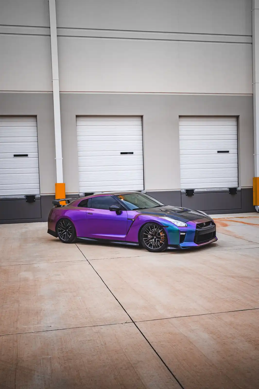 A Nissan GT-R sports car with a vibrant purple and blue iridescent paint job is parked on a concrete lot in front of a…
