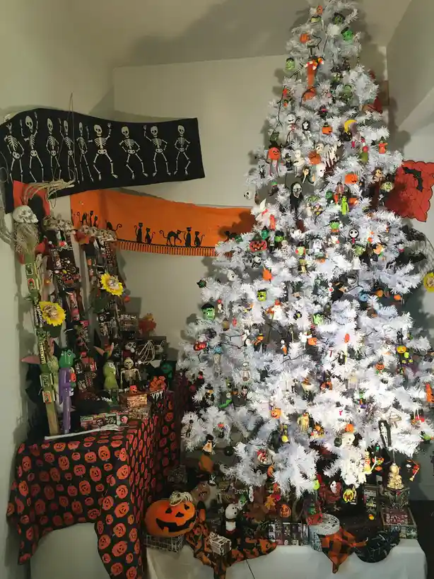 A white artificial Christmas tree is densely decorated with numerous Halloween-themed ornaments, including pumpkins…
