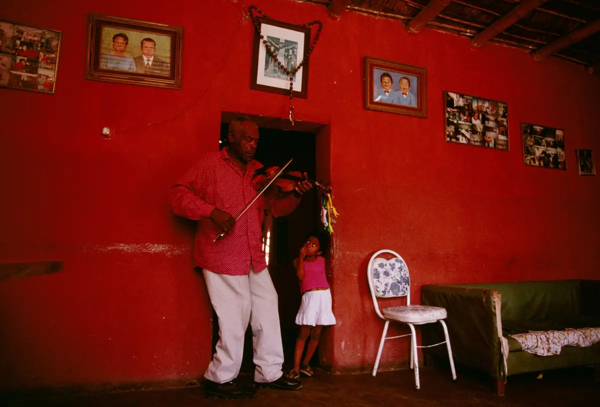 A man in a red shirt and white pants stands in a doorway playing a violin, while a small child in a pink top and white skirt…