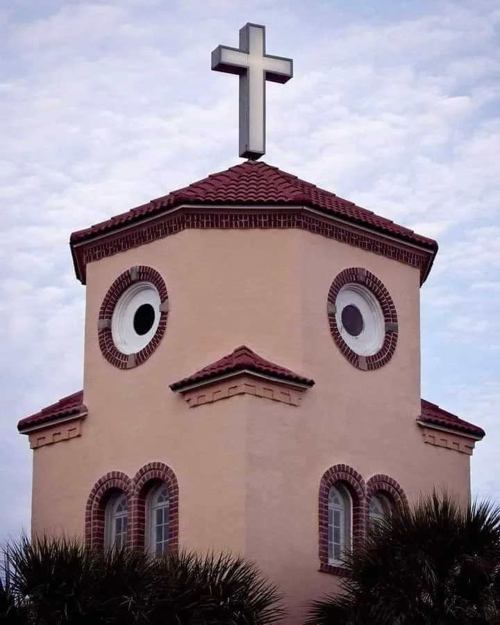 A tan church steeple with a red-tiled roof and a silver cross on top, has two large circular windows that resemble eyes…