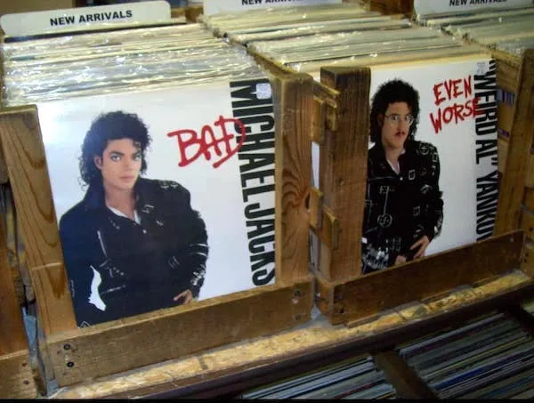 Two adjacent vinyl record sleeves are displayed side-by-side in a wooden rack. The left sleeve features Michael Jackson's…