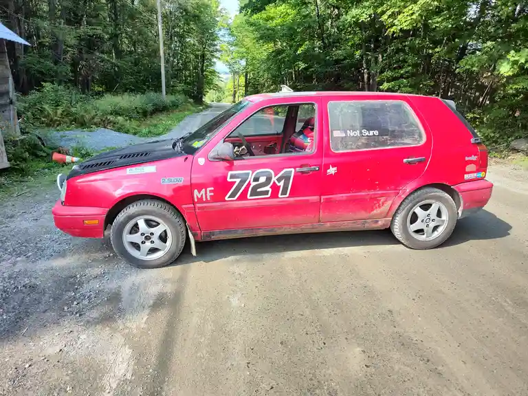 A red, modified Volkswagen Golf rally car, covered in dirt and bearing the number 721, is parked on a gravel road surrounded…