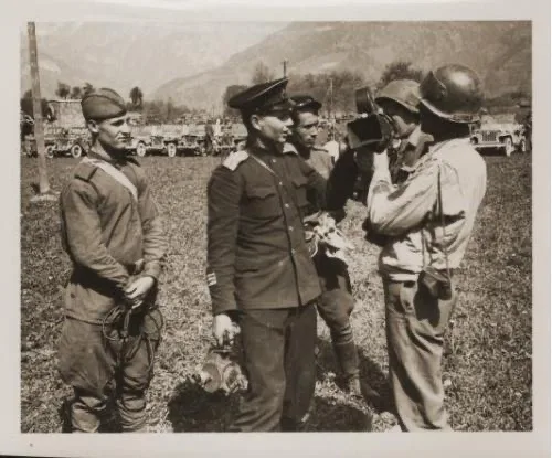 A black-and-white historical photo shows four men in military uniforms standing outdoors near vehicles and mountains. One…