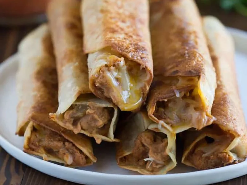 A close-up photo shows a pile of golden-brown, crispy taquitos on a white plate, with melted cheese and filling visibly…