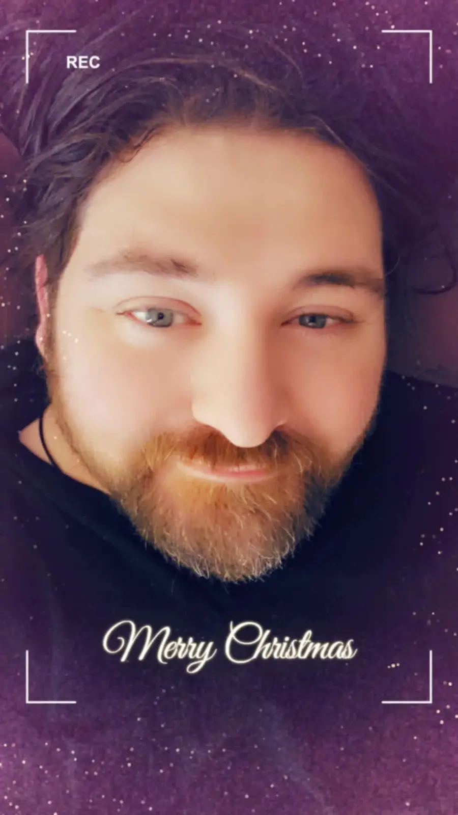A selfie of a man with a beard and long hair, looking at the camera with a slight smile, set against a purple starry…