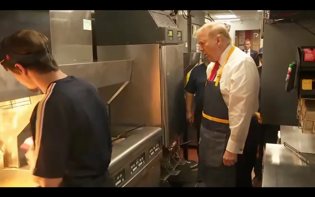 A man in a white shirt, red tie, and dark apron with yellow straps stands in a commercial kitchen, looking intently at…