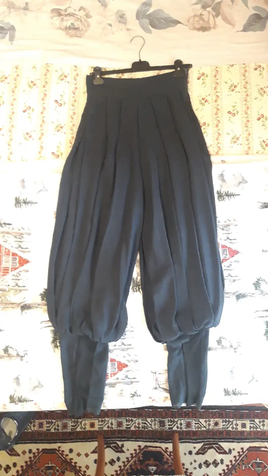 A pair of dark blue, extremely baggy pants hangs on a black hanger against a patterned wallpaper background. The pants have…