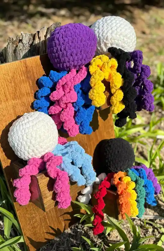 Several colorful, crocheted octopus toys with yarn bodies and tentacles are displayed on wooden blocks outdoors. The…
