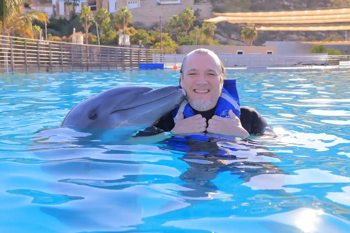 A man with a white beard and blue life vest smiles broadly while in a bright blue pool, being affectionately nuzzled by a…