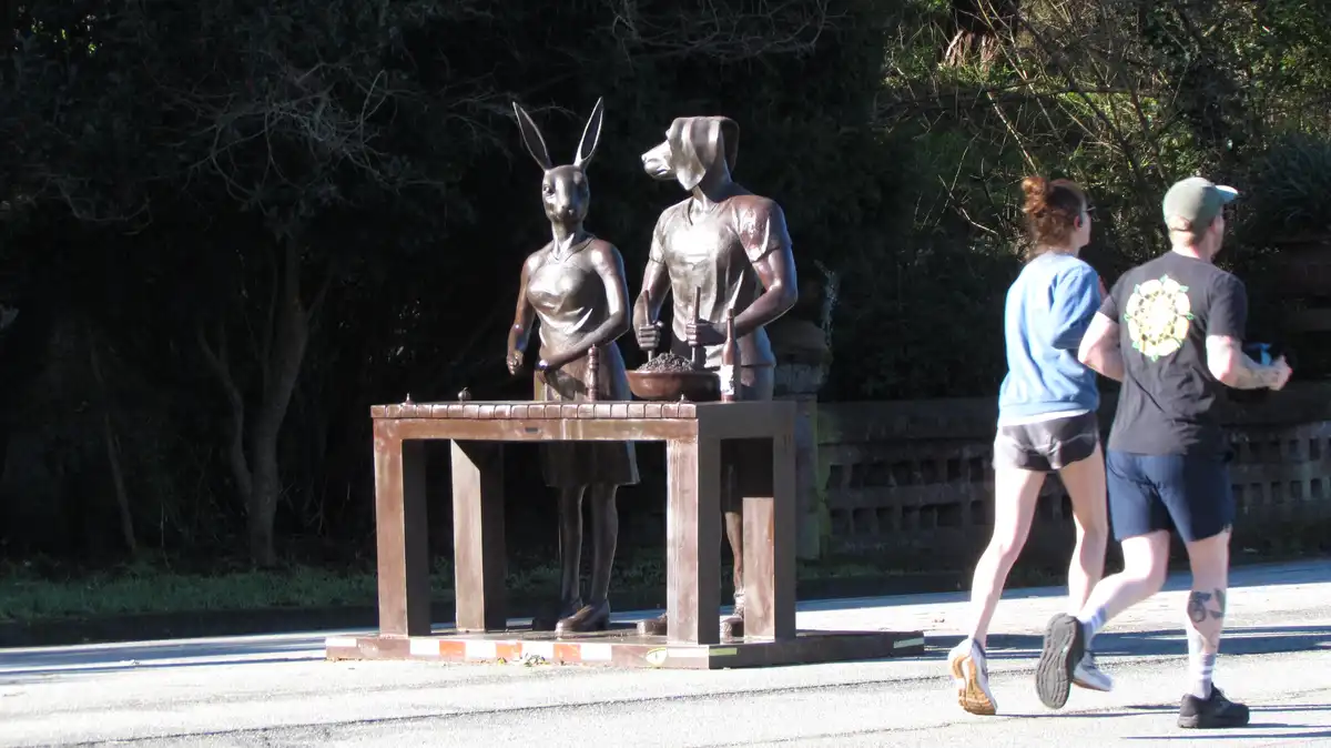 Two bronze statues of a kangaroo and a dog, both wearing human clothing and standing at a table with a bowl and bottles, are…
