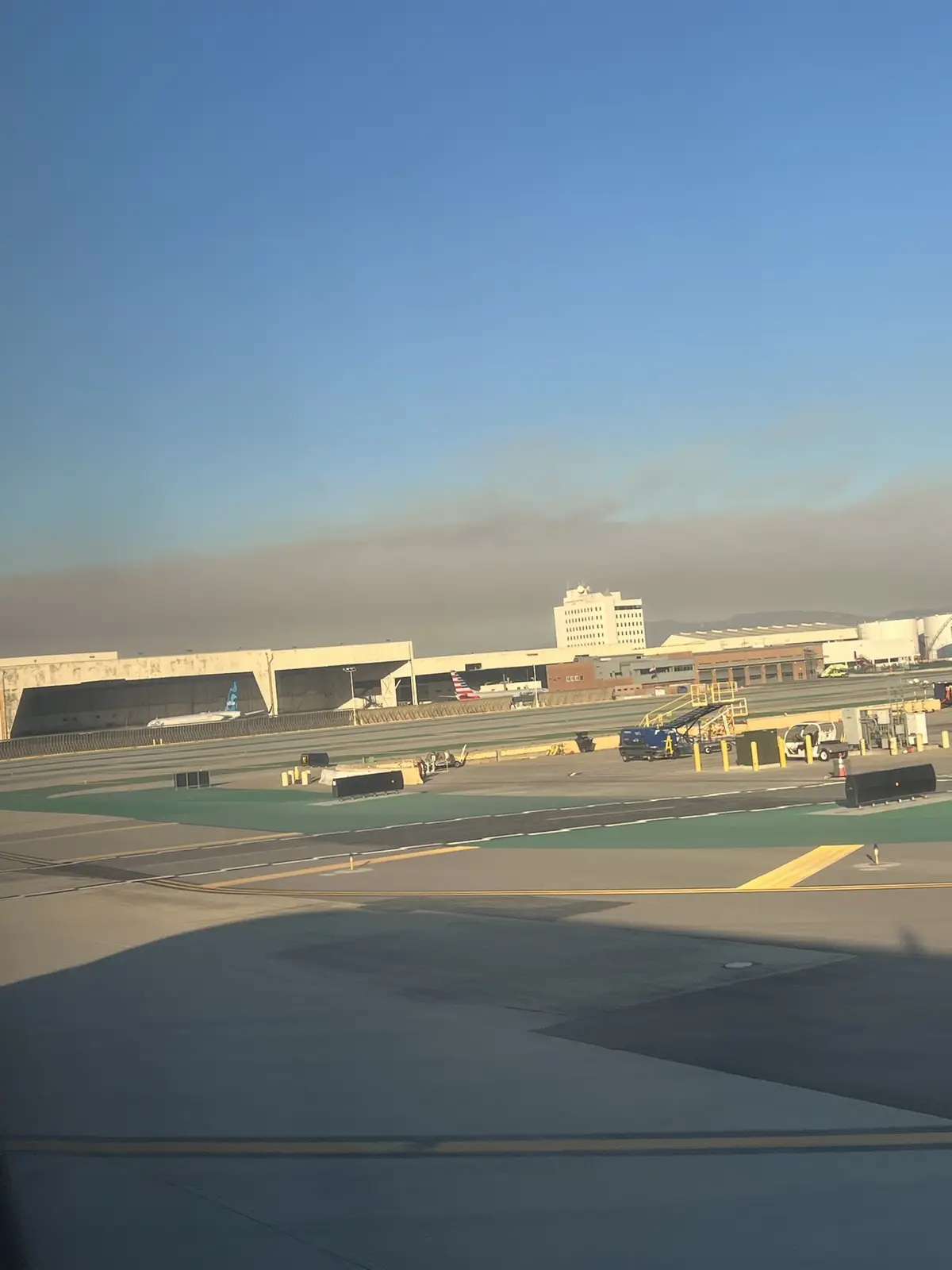 An airport tarmac scene viewed from an airplane window, showing runways, terminal buildings, parked aircraft, and ground…