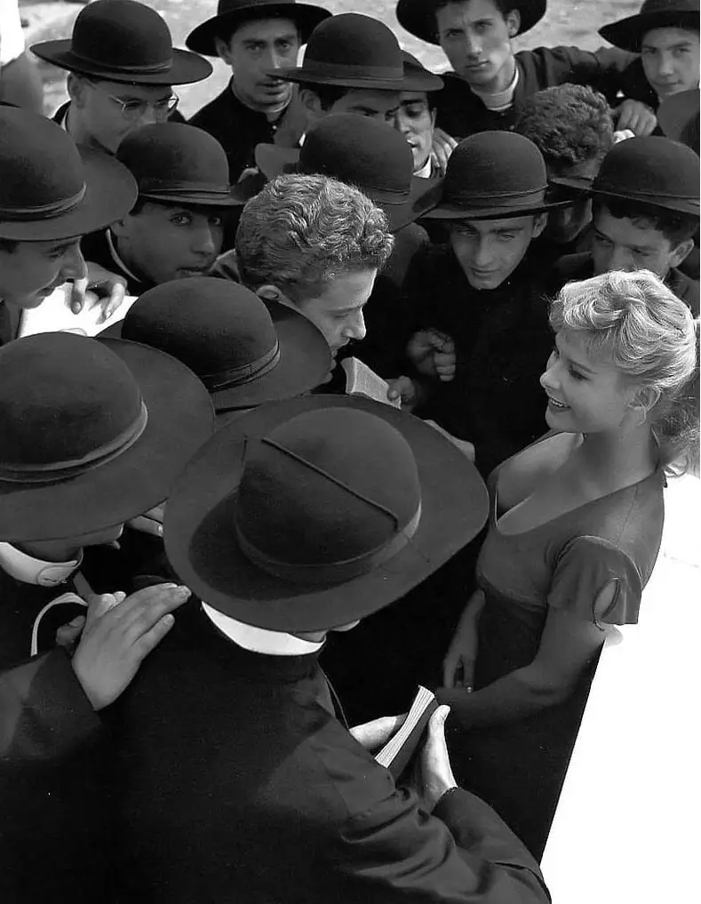 a black-and-white photo shows a woman in a dress smiling while surrounded by men in dark robes and bowler hats, one of whom…