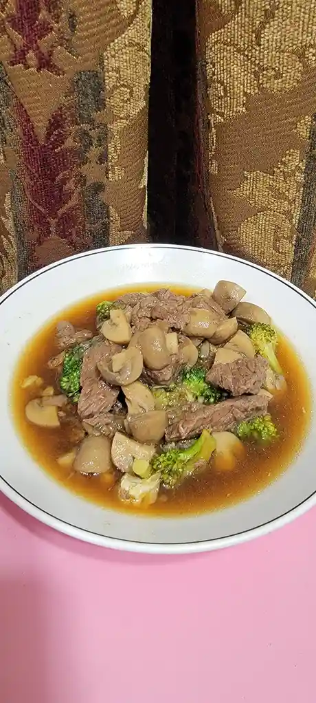 A white bowl with a black rim sits on a pink surface, filled with a brown broth containing chunks of beef, sliced mushrooms…