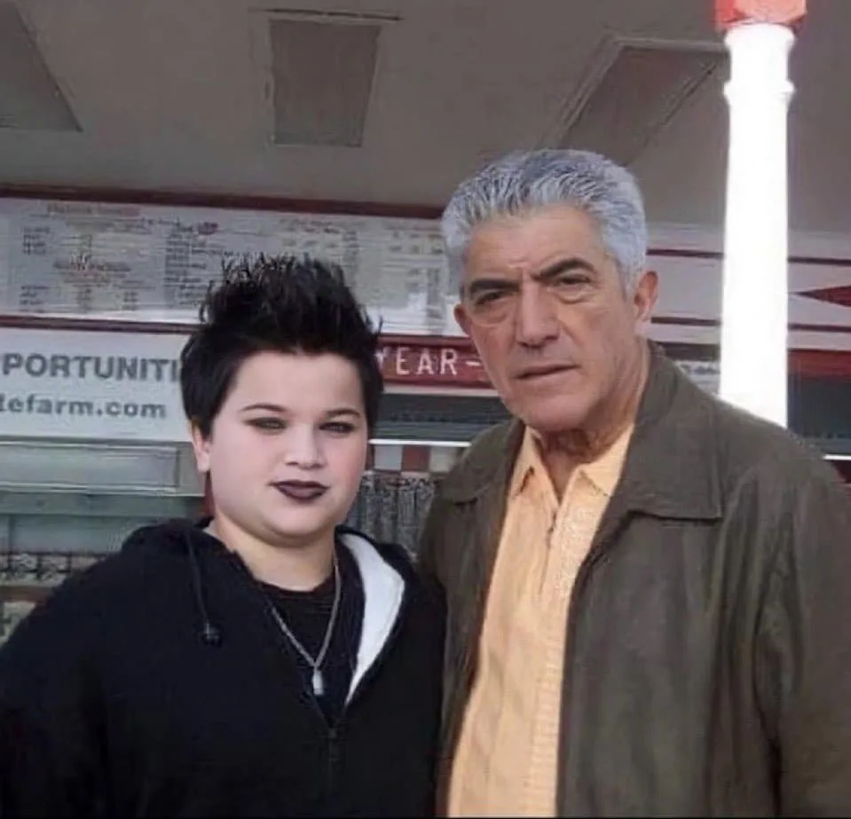 A young person with dark lipstick and spiky hair stands beside an older man with gray hair and a stern expression, both…