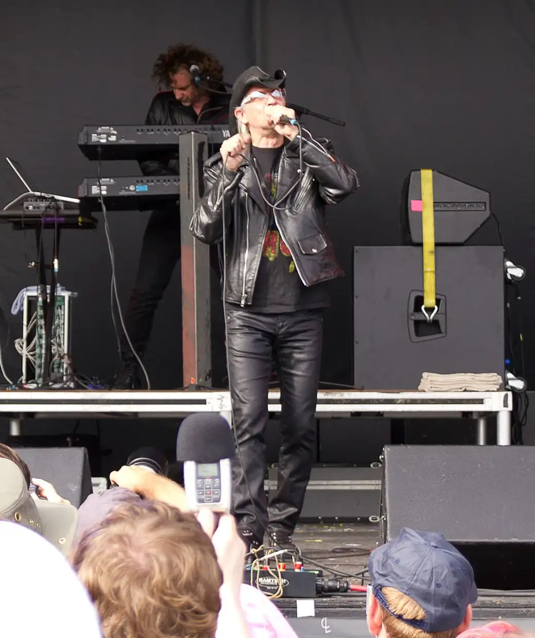 A man in a black leather jacket and hat sings into a microphone on a stage, with a keyboardist behind him. In the…