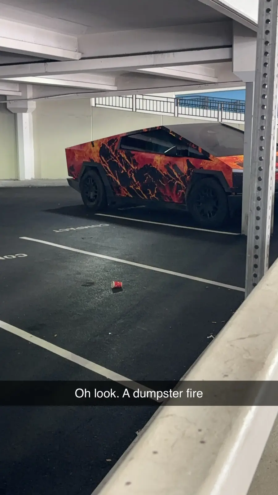 A Tesla Cybertruck with a fiery lava-like wrap is parked in an underground parking garage, partially obscured by a metal…