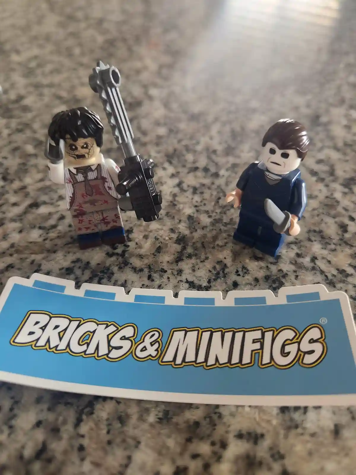Two LEGO minifigures stand on a speckled countertop; one is a zombie-like figure with a blood-stained shirt holding a large…