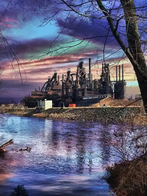 A photograph shows a large, rusted steel mill complex with numerous smokestacks and pipes, situated beside a river under a…