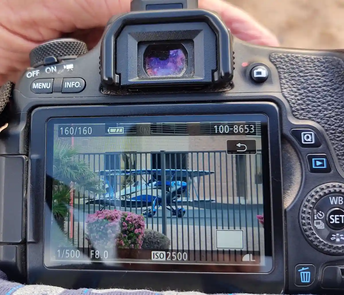 A close-up of a DSLR camera's top and rear, showing a hand holding it. The camera's viewfinder displays a colorful, abstract…