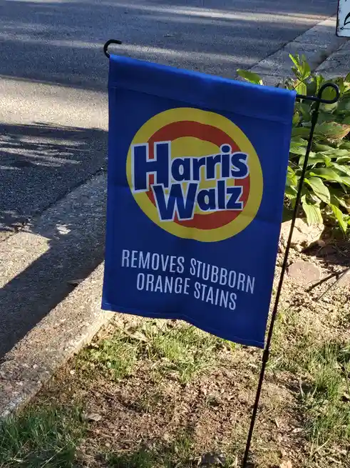 A blue yard flag with the logo and text 'Harris Walz REMOVES STUBBORN ORANGE STAINS' is stuck in grass next to a sidewalk…