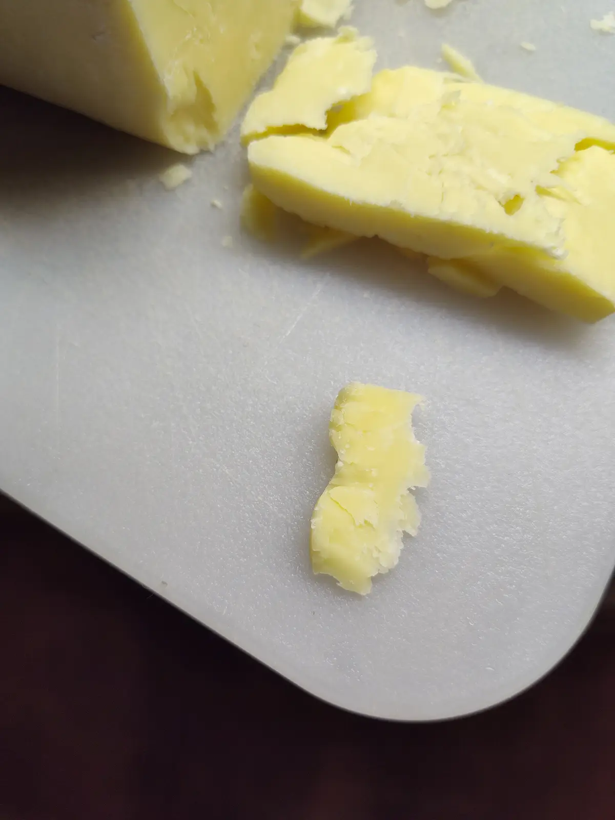 A close-up photo shows a small, irregularly shaped piece of yellow butter on a white cutting board, with larger chunks of…