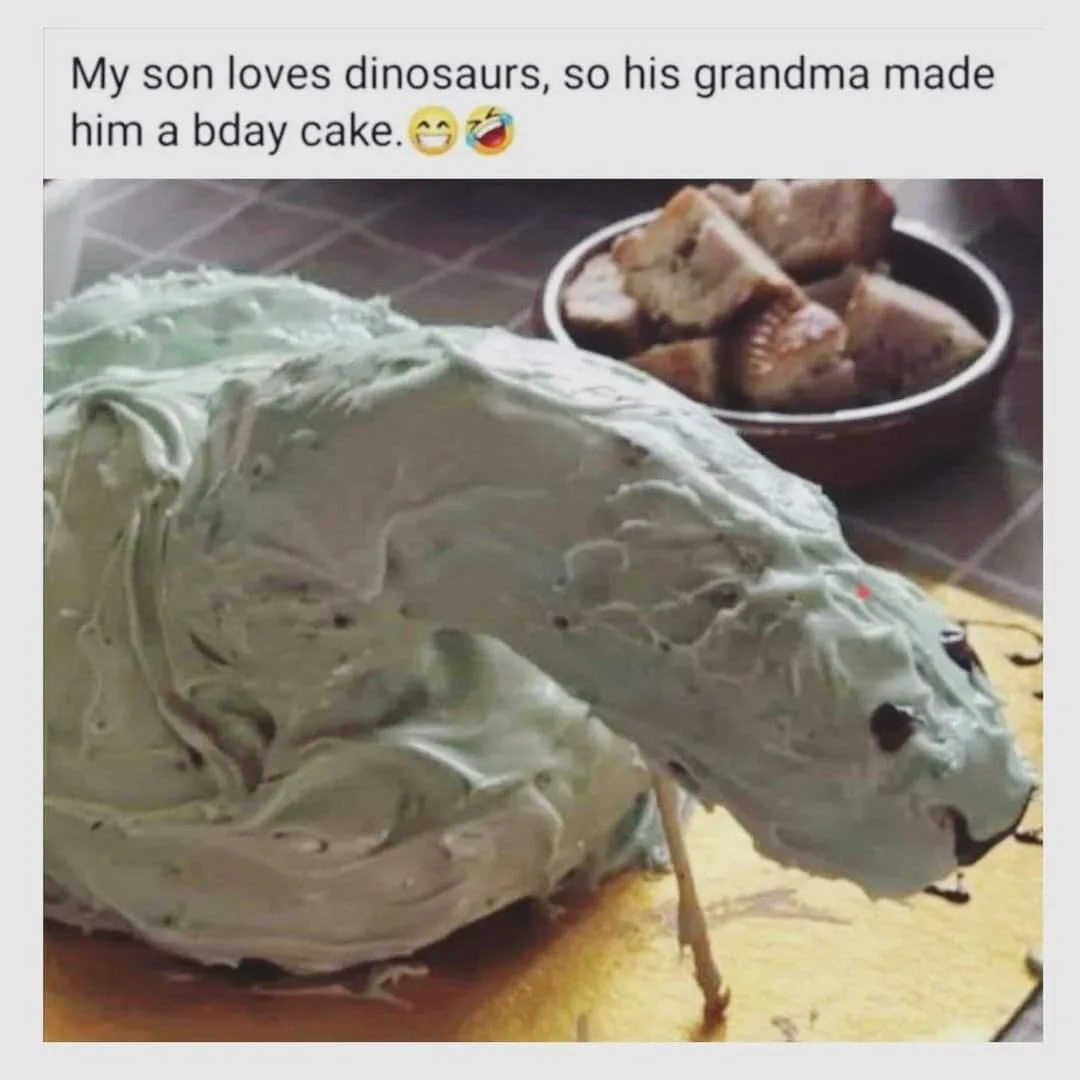 A poorly decorated birthday cake shaped like a dinosaur, covered in lumpy, light green frosting with two small dark eyes and…