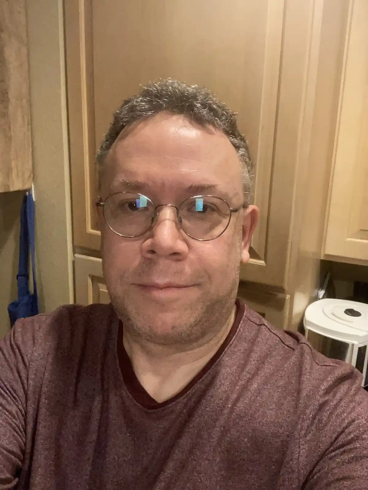 A man with glasses and a maroon shirt smiles slightly at the camera in a kitchen with wooden cabinets. The overlaid text…