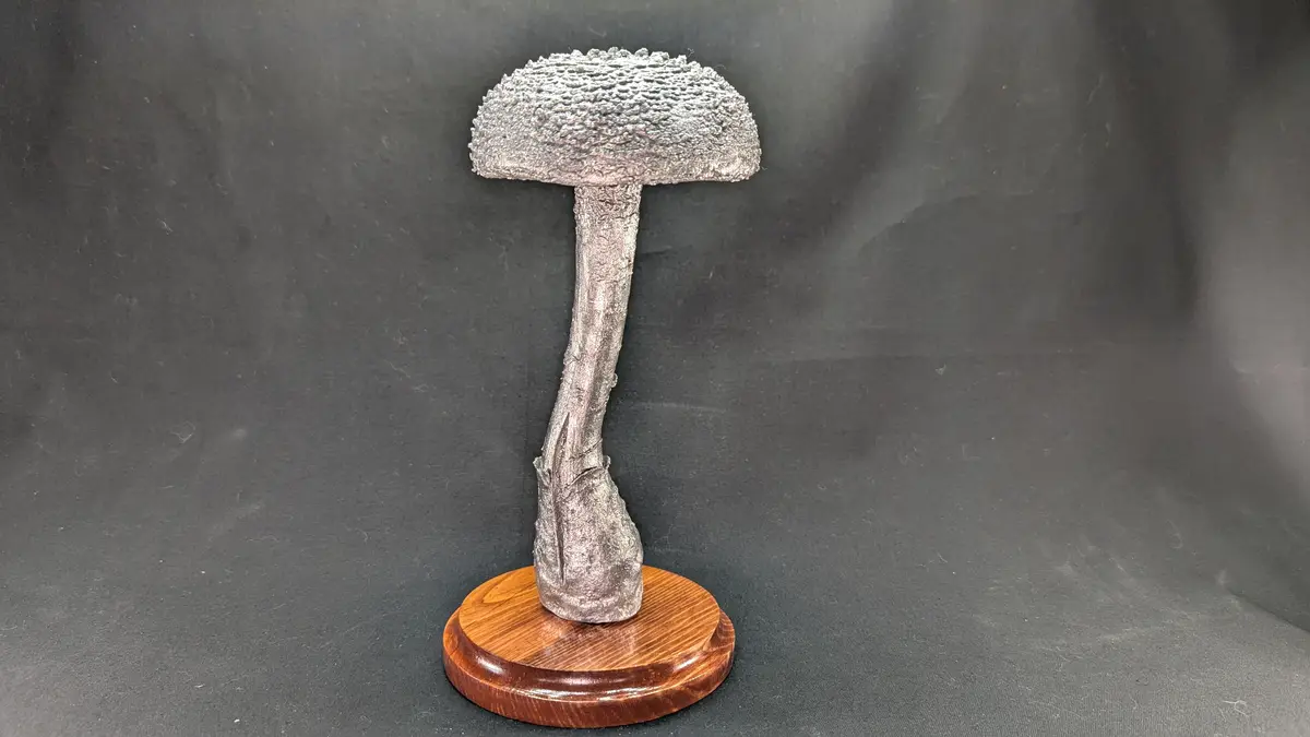 A silver-colored, textured mushroom sculpture with a curved stem stands on a polished wooden circular base against a dark…