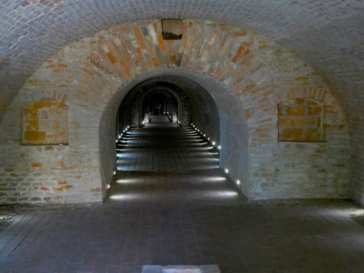 A dimly lit, arched brick tunnel with a long staircase receding into the distance, illuminated by small lights along the…