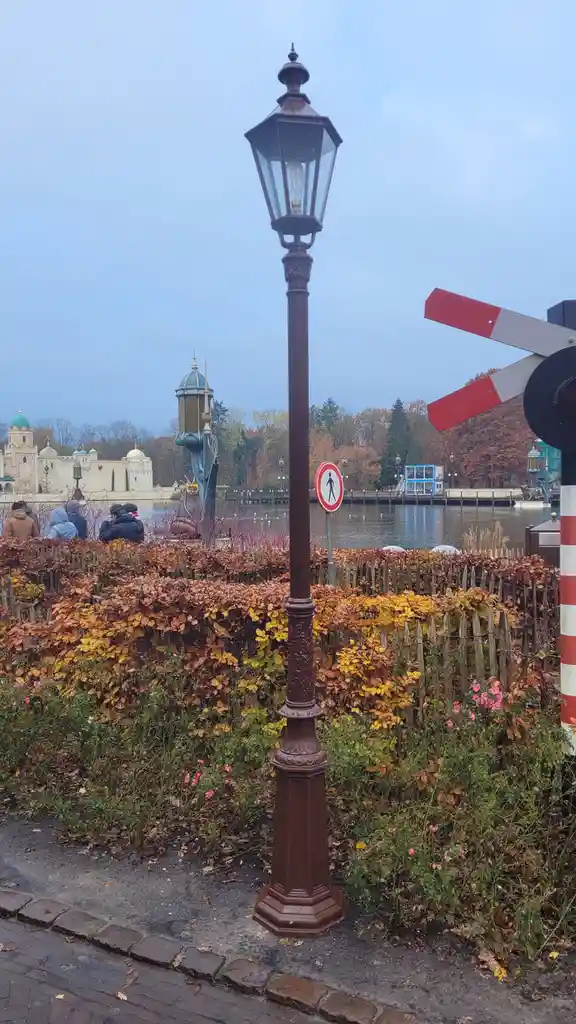 A photo of a vintage-style brown lamppost standing on a cobblestone path next to autumn-colored bushes and a wooden fence…