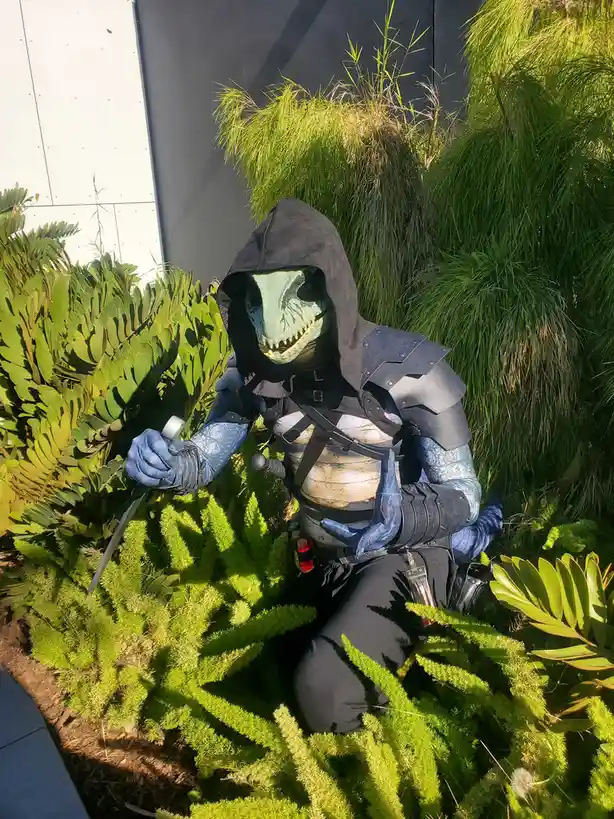 A person in a detailed costume of a reptilian humanoid with a green scaly face, black hood, and armor is crouching among…