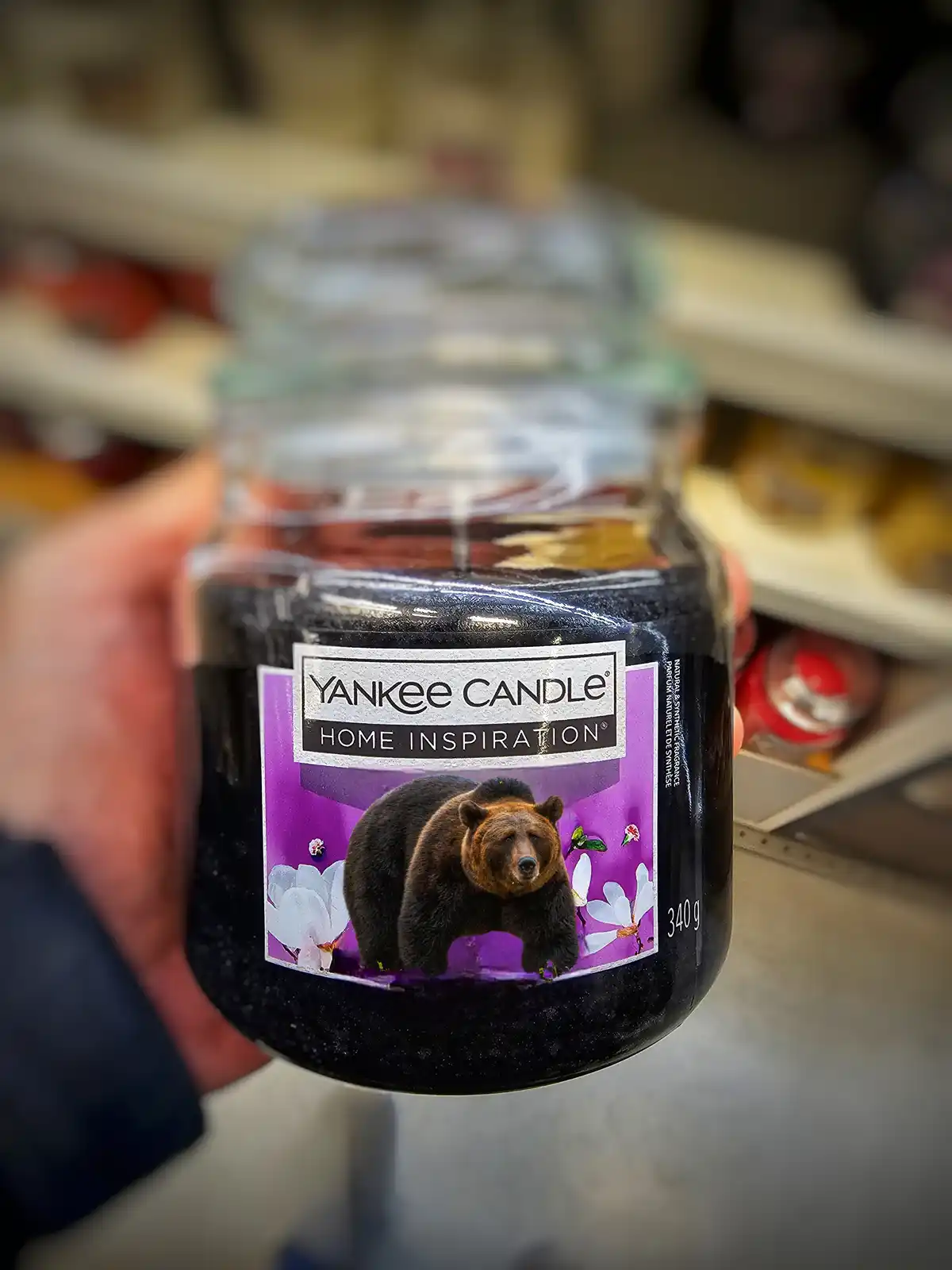 A hand holds a Yankee Candle jar labeled 'Home Inspiration' with a label featuring a brown bear standing among white flowers…