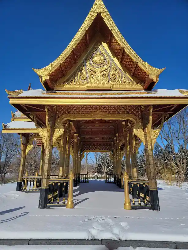 A golden, ornate Thai-style temple structure with intricate carvings and red accents stands covered in snow under a clear…