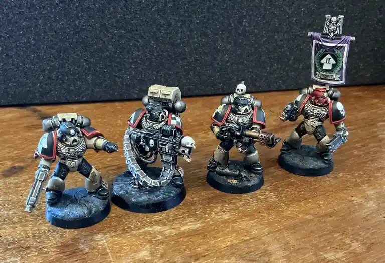 Four painted miniature figurines of Space Marines in power armor stand on a wooden surface against a dark background. The…
