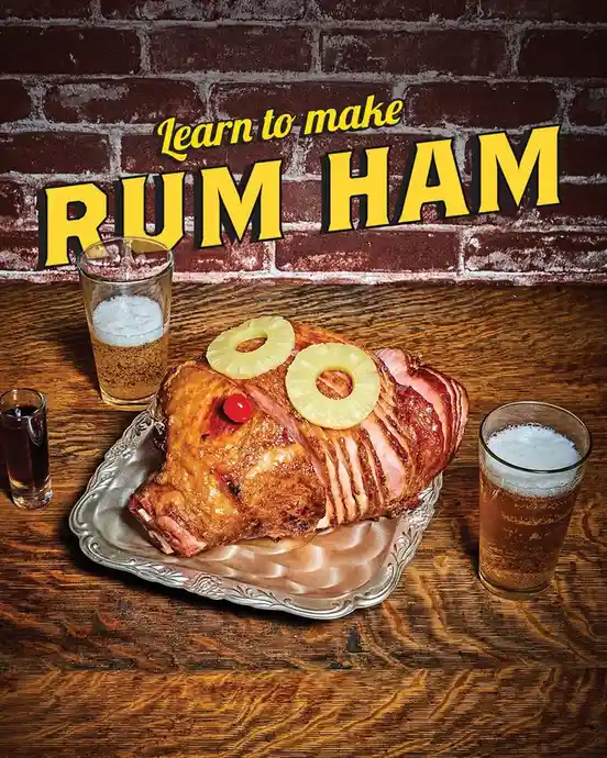 A large, glazed ham sits on a silver platter on a wooden table, decorated with two pineapple rings and a cherry on top…