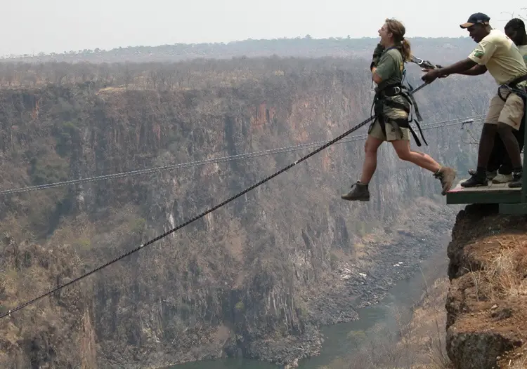 A woman in safari gear is mid-stride on a narrow rope bridge spanning a deep canyon, looking upward with a fearful…